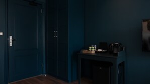 A dark blue room with a wooden floor, a door with a handle, a small table with a kettle and cups, and a framed picture on the wall.