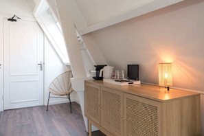 A room with a sloped ceiling, a white door, a wicker chair, a wooden cabinet with a woven front, and a table with a lamp.