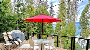 Een houten terras met een tafel en stoelen, een rode parasol en uitzicht op bomen en een waterpartij.