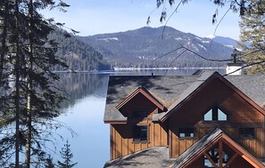 Une cabane au revêtement extérieur en bois, avec un toit en pente, et une vue sur un lac paisible et des montagnes en arrière-plan.