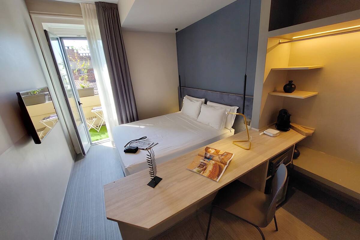 A hotel room with a bed, bedside table, desk, chair, and a small shelf with a vase.