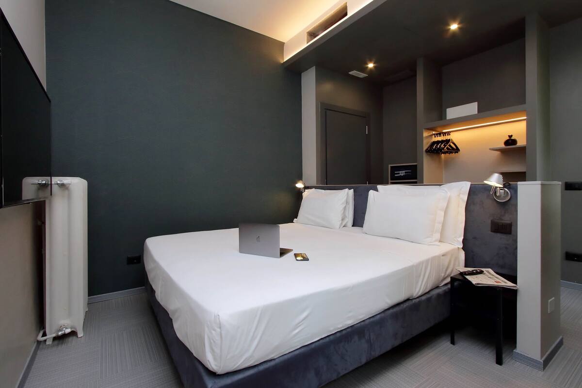A modern hotel room with a large bed, a desk, and a chair. There is a mirror above the bed and a shelf with a lamp.