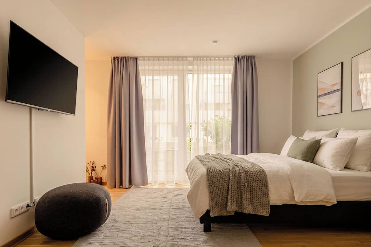 Un dormitorio moderno con cama, televisor de pantalla plana, ventana grande con cortinas y un pequeño otoman redondo.