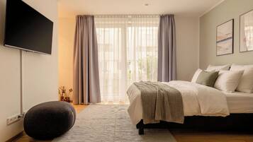Un dormitorio moderno con cama, televisor de pantalla plana, ventana grande con cortinas y un pequeño otoman redondo.