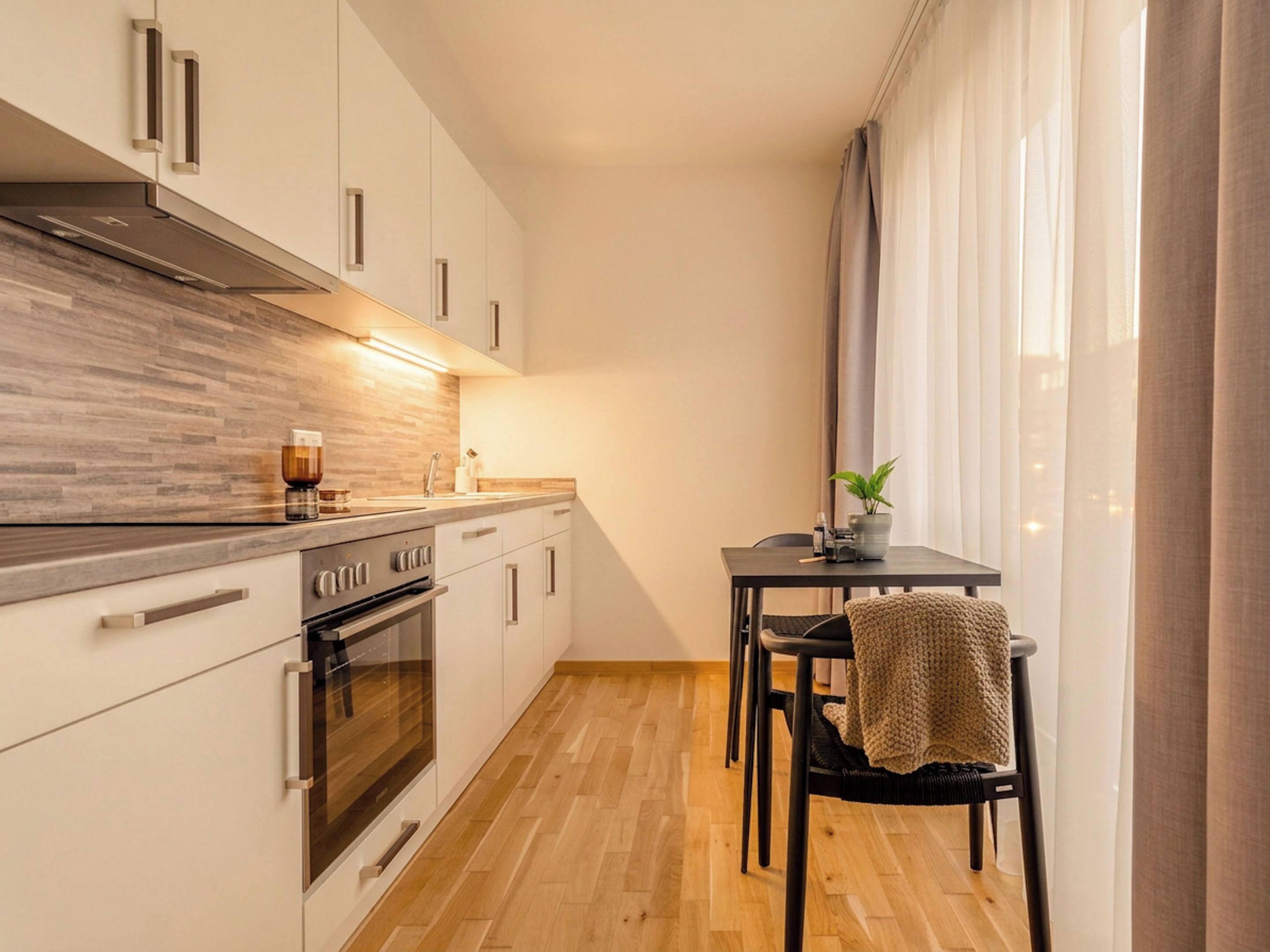 A modern kitchen with a built-in oven, a dining area with a table and chairs, and a large window with curtains.