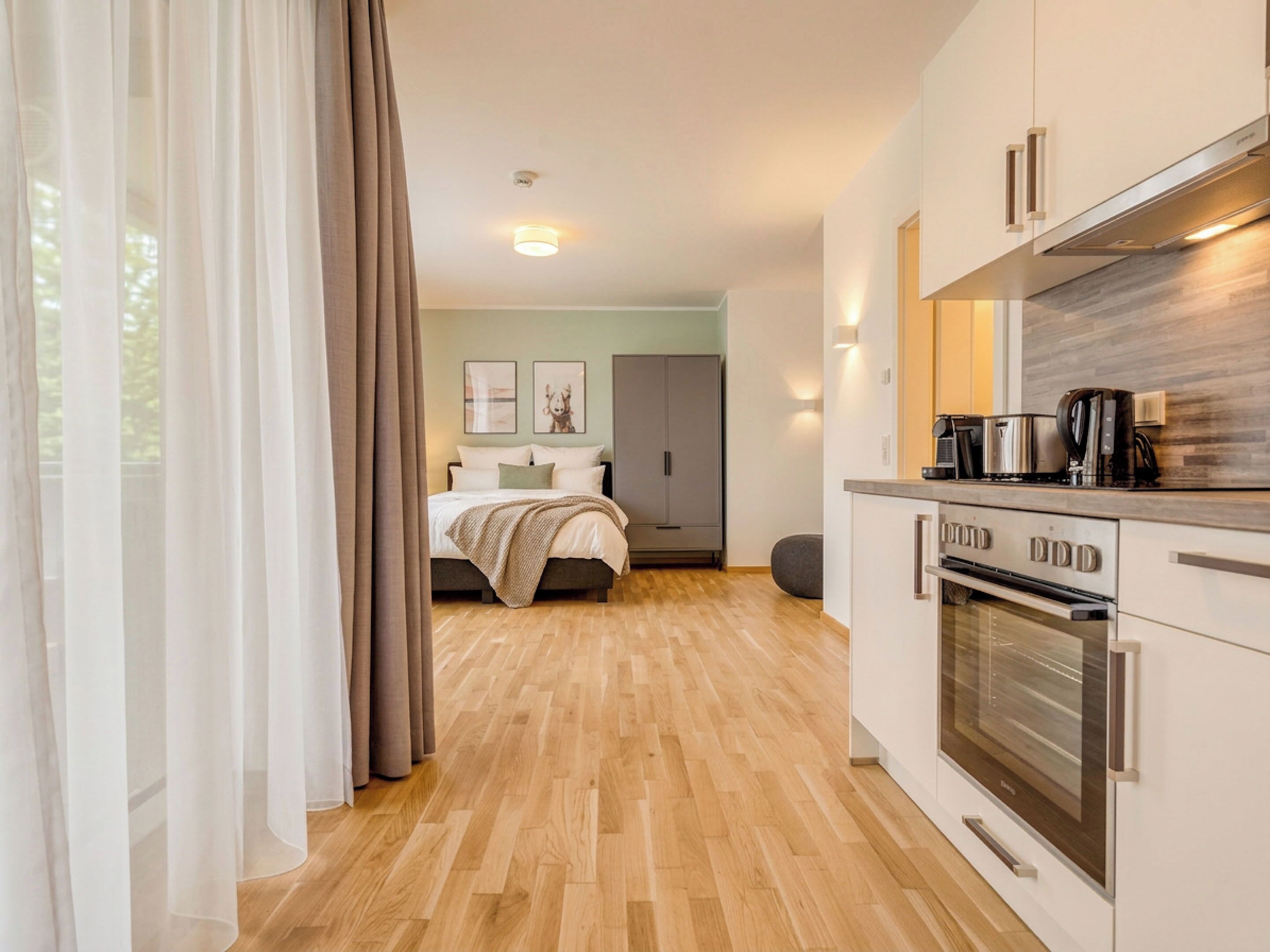 A modern hotel room with a wooden floor, a bed with a beige blanket, a kitchenette with a stove and coffee maker, and a view of the outside through sheer curtains.