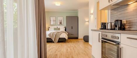 A modern hotel room with a wooden floor, a bed with a beige blanket, a kitchenette with a stove and coffee maker, and a view of the outside through sheer curtains.