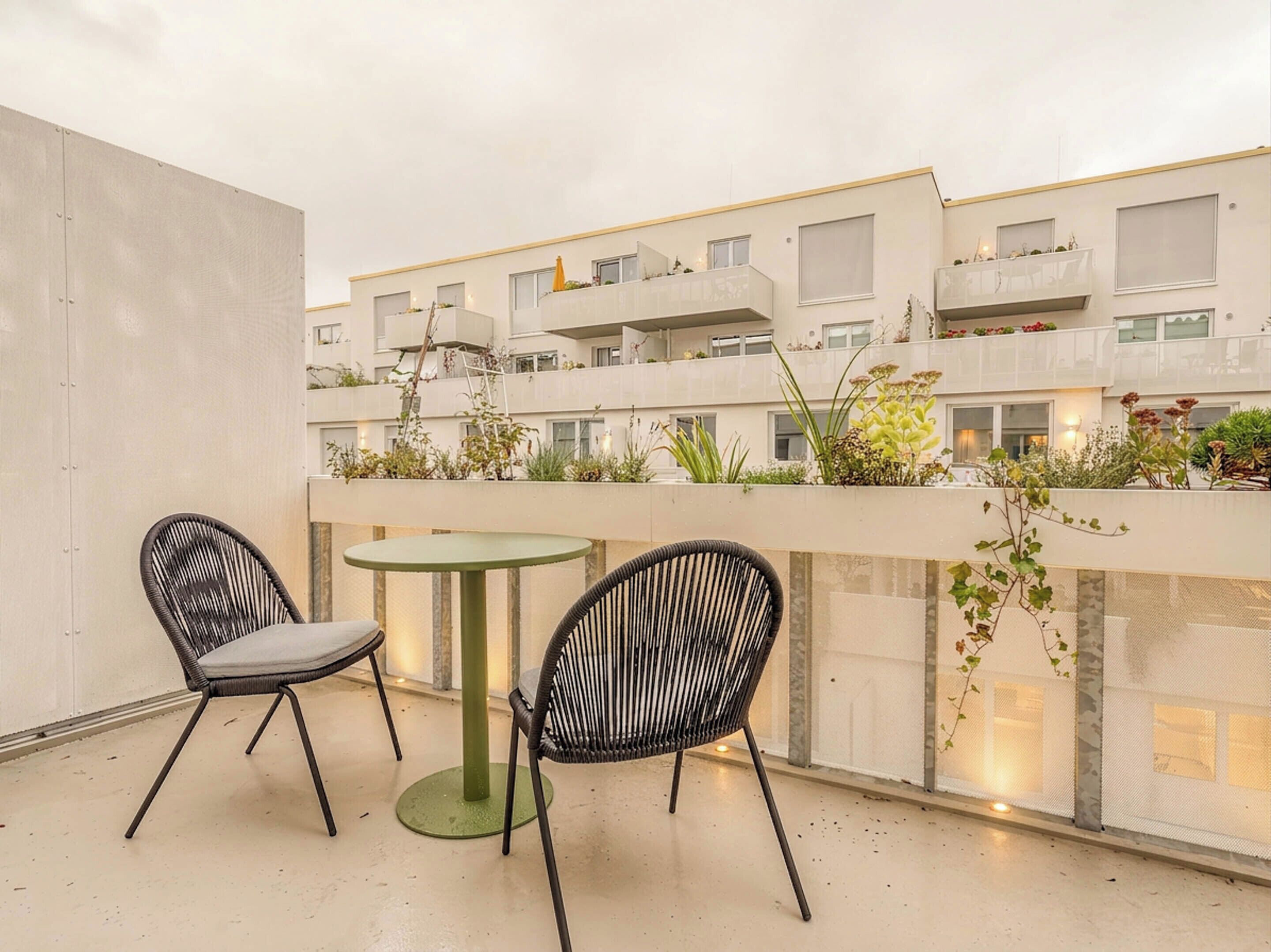 Un balcón con dos sillas y una mesa, con vista a un edificio con balcones y plantas.