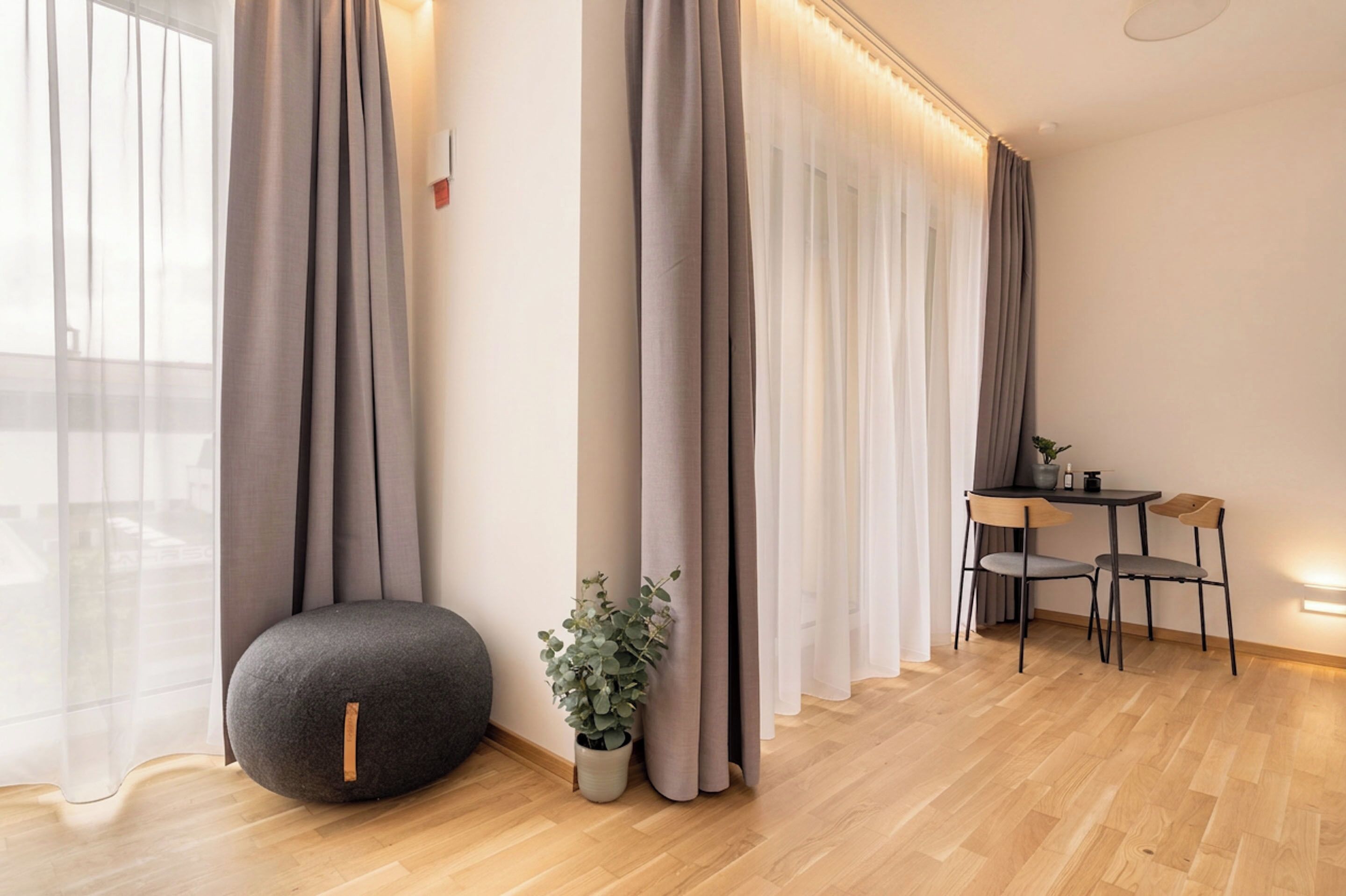 A modern room with a wooden floor, a round ottoman, a small table with chairs, and sheer curtains.