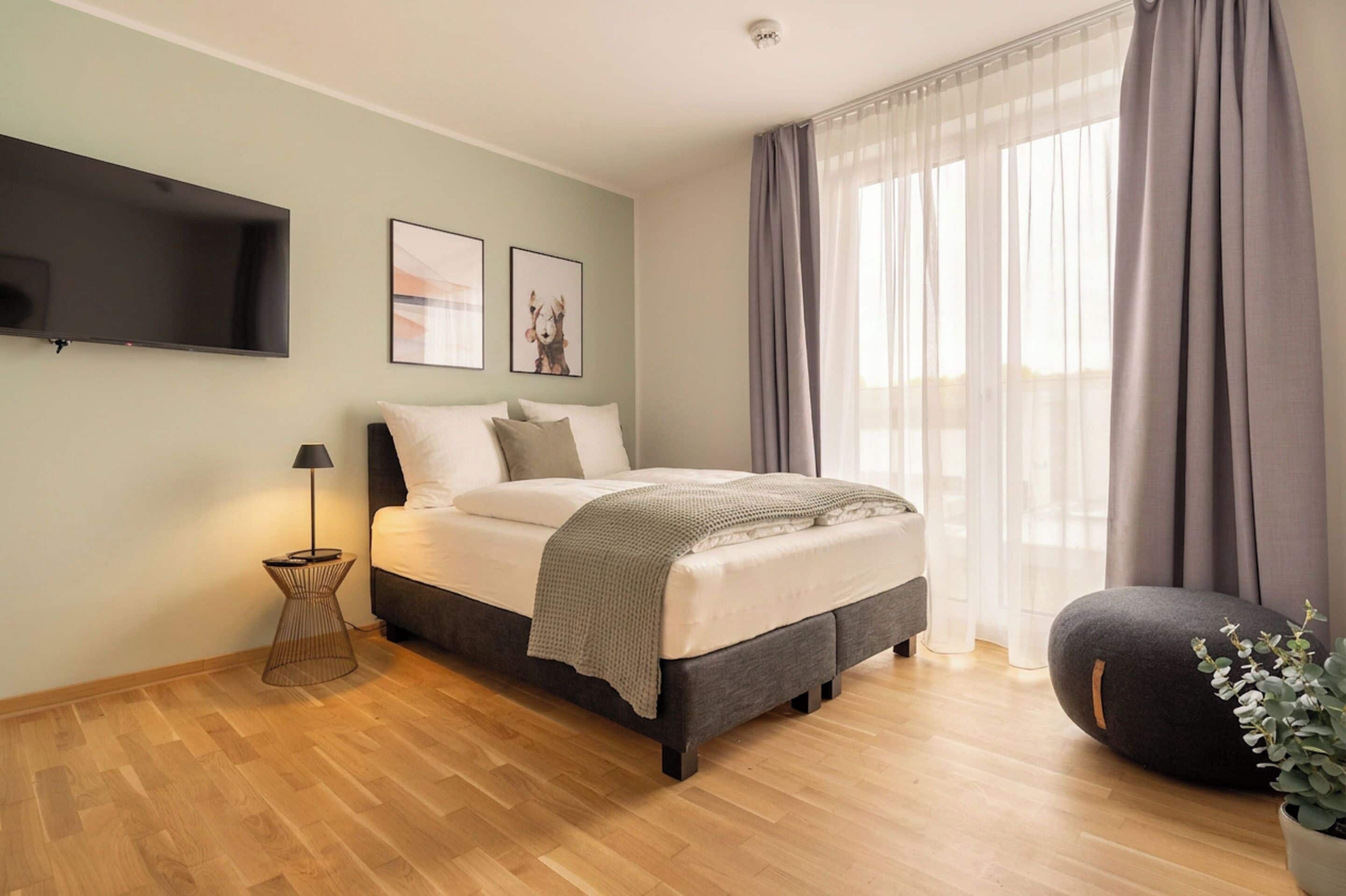 Un dormitorio con una cama, televisor, mesita de noche y ventana con cortinas.