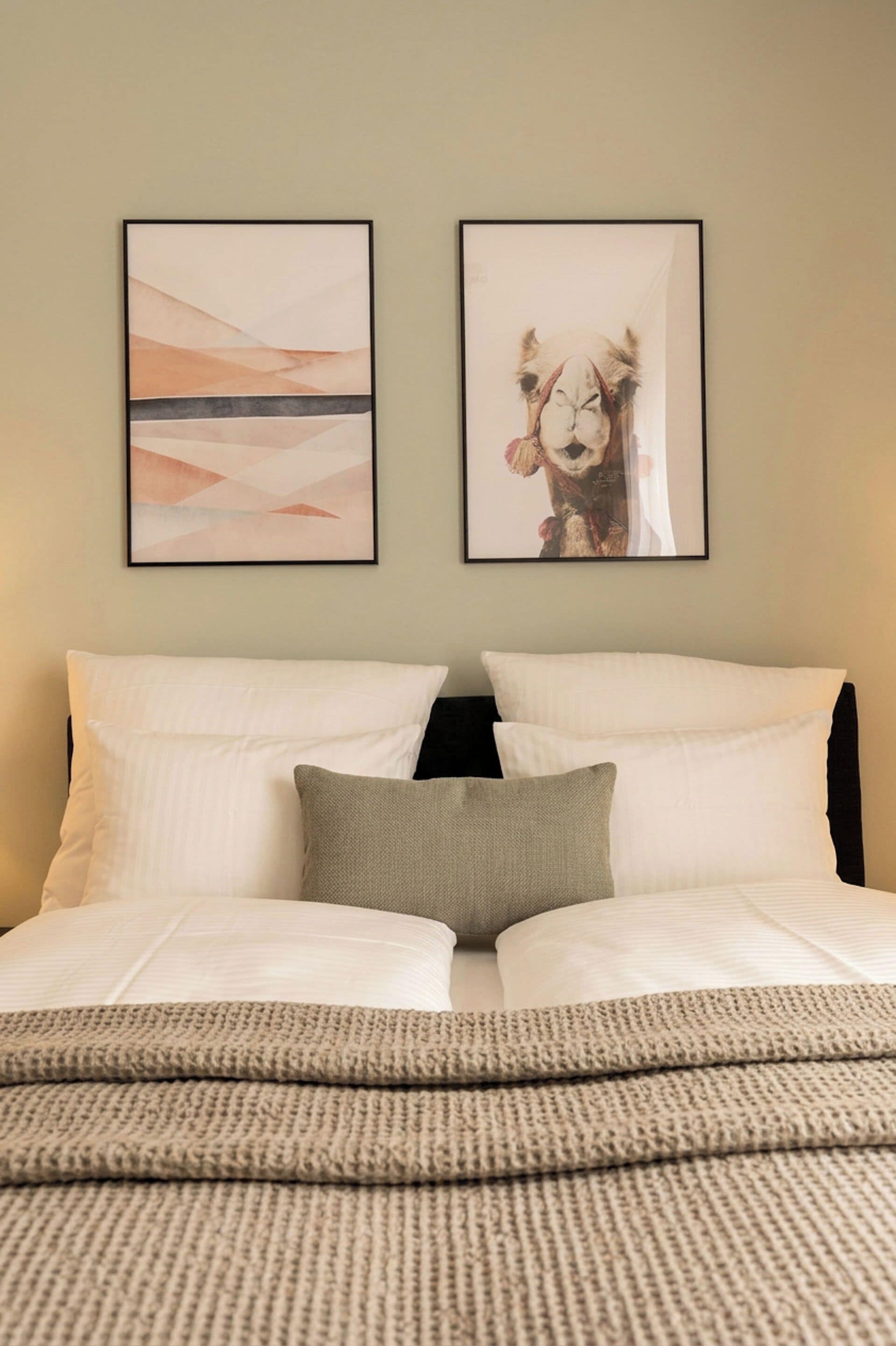 Ein Bett mit weißer Bettwäsche und einem grauen Kopfkissen, zwei gerahmte Kunstwerke an der Wand und eine strukturierte, beige Decke.