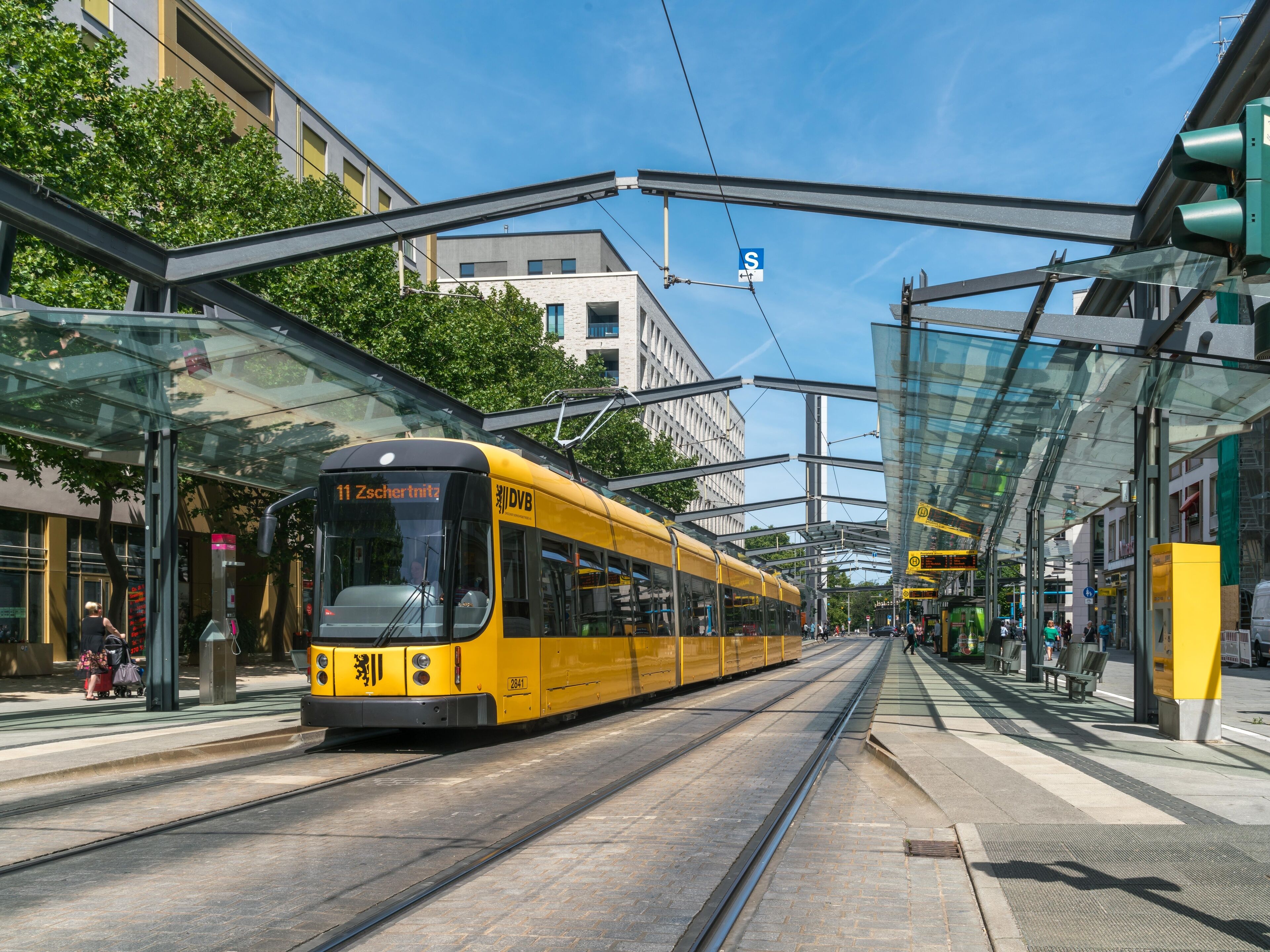 Un tramway jaune se trouve dans une station de tramway moderne, dotée d’abris en verre, sous un ciel dégagé.