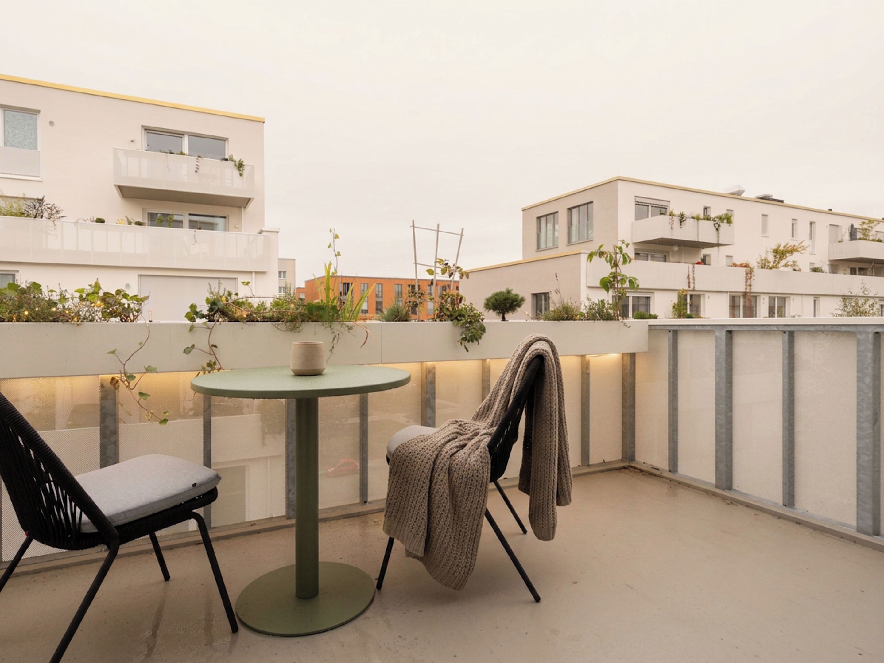 En balkon med et rundt bord, to stole, et strikket tæppe og plantekrukker.