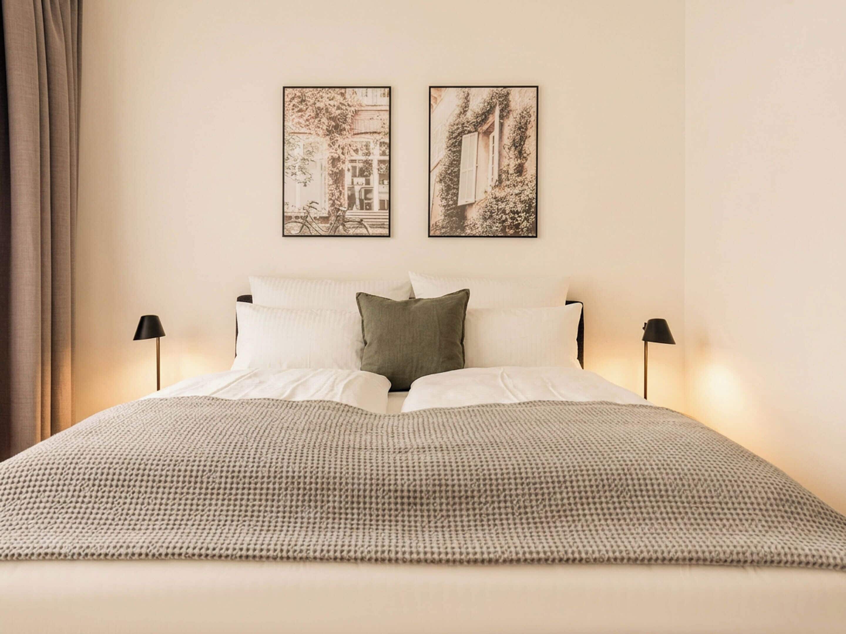 A neatly made bed with a patterned bedspread, two wall-mounted pictures, and two bedside lamps.