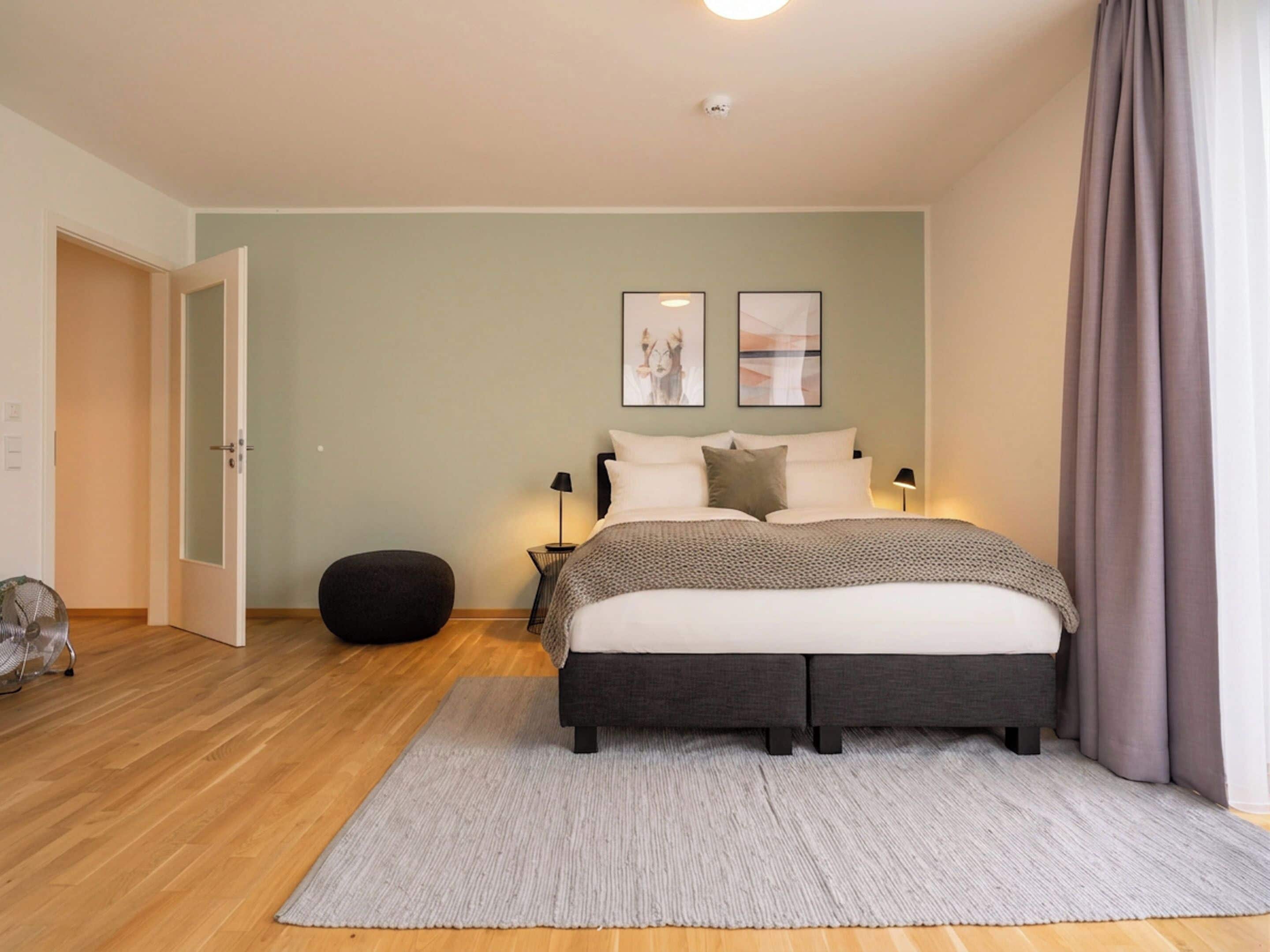 A bedroom with a bed, bedside lamps, a rug, a black ottoman, and a door.