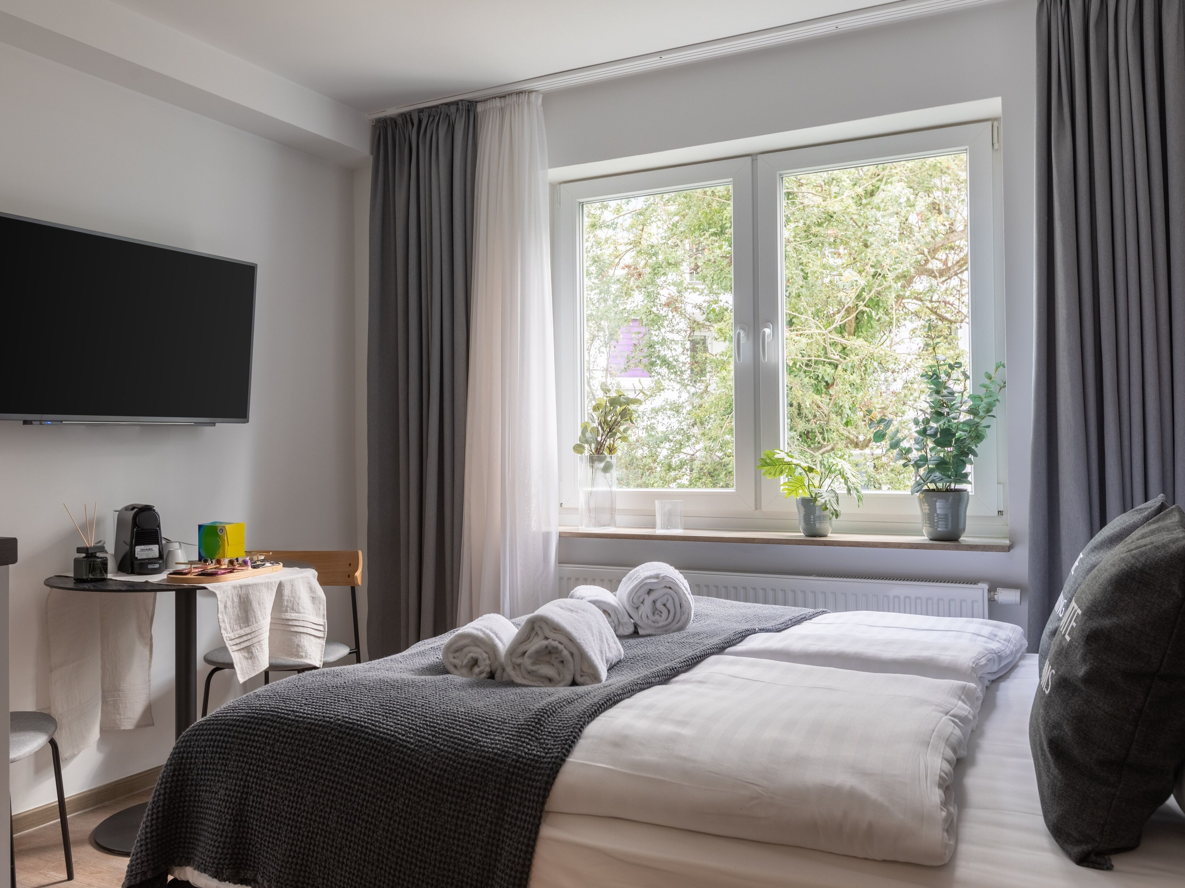 A hotel room with a bed, a flat-screen TV, a small table with a coffee maker, and a window with plants.