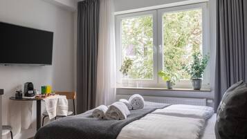 A hotel room with a bed, a flat-screen TV, a small table with a coffee maker, and a window with plants.