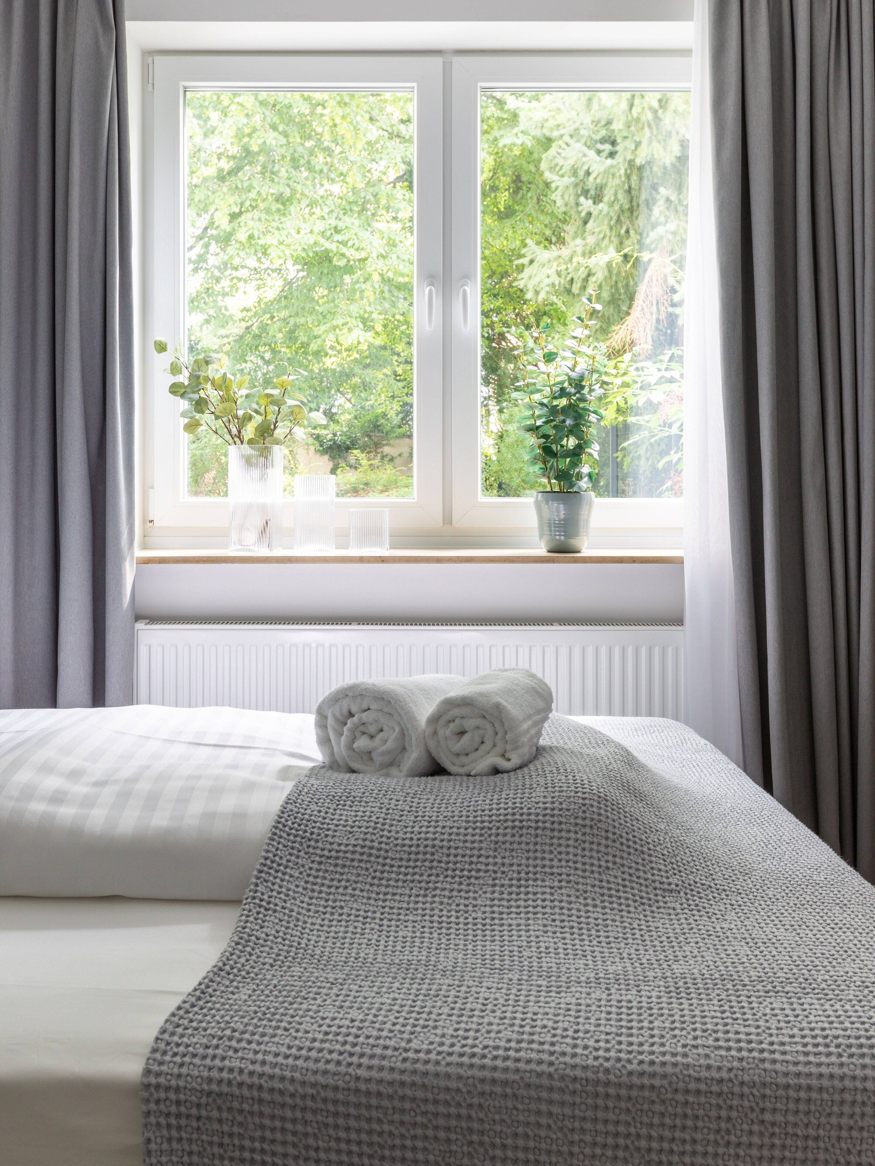 A bedroom with a bed, a window with curtains, and two potted plants on the windowsill.