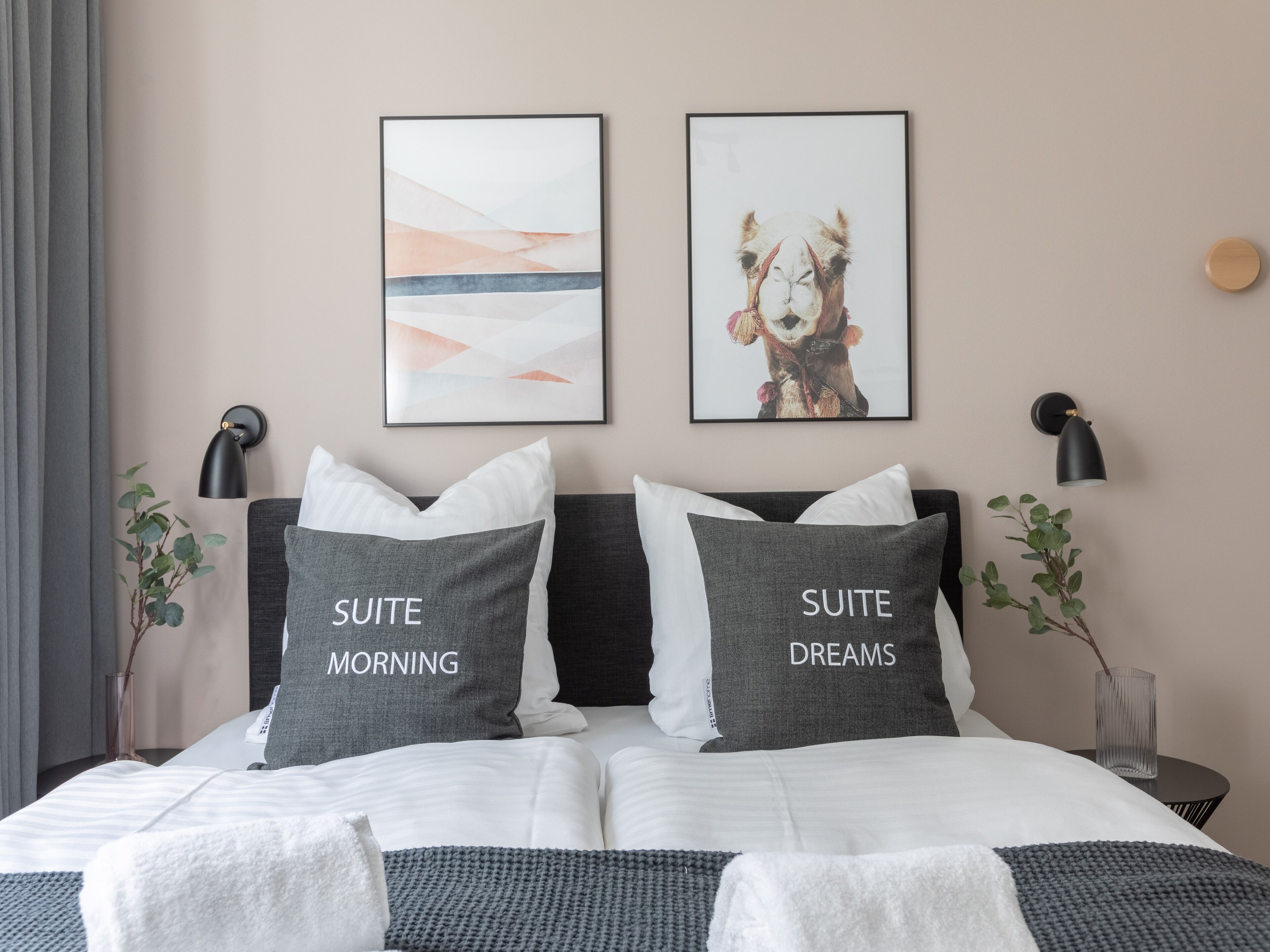 A neatly made bed with two pillows labeled "SUITE MORNING" and "SUITE DREAMS," a wall-mounted abstract painting, and a framed photo of a camel.