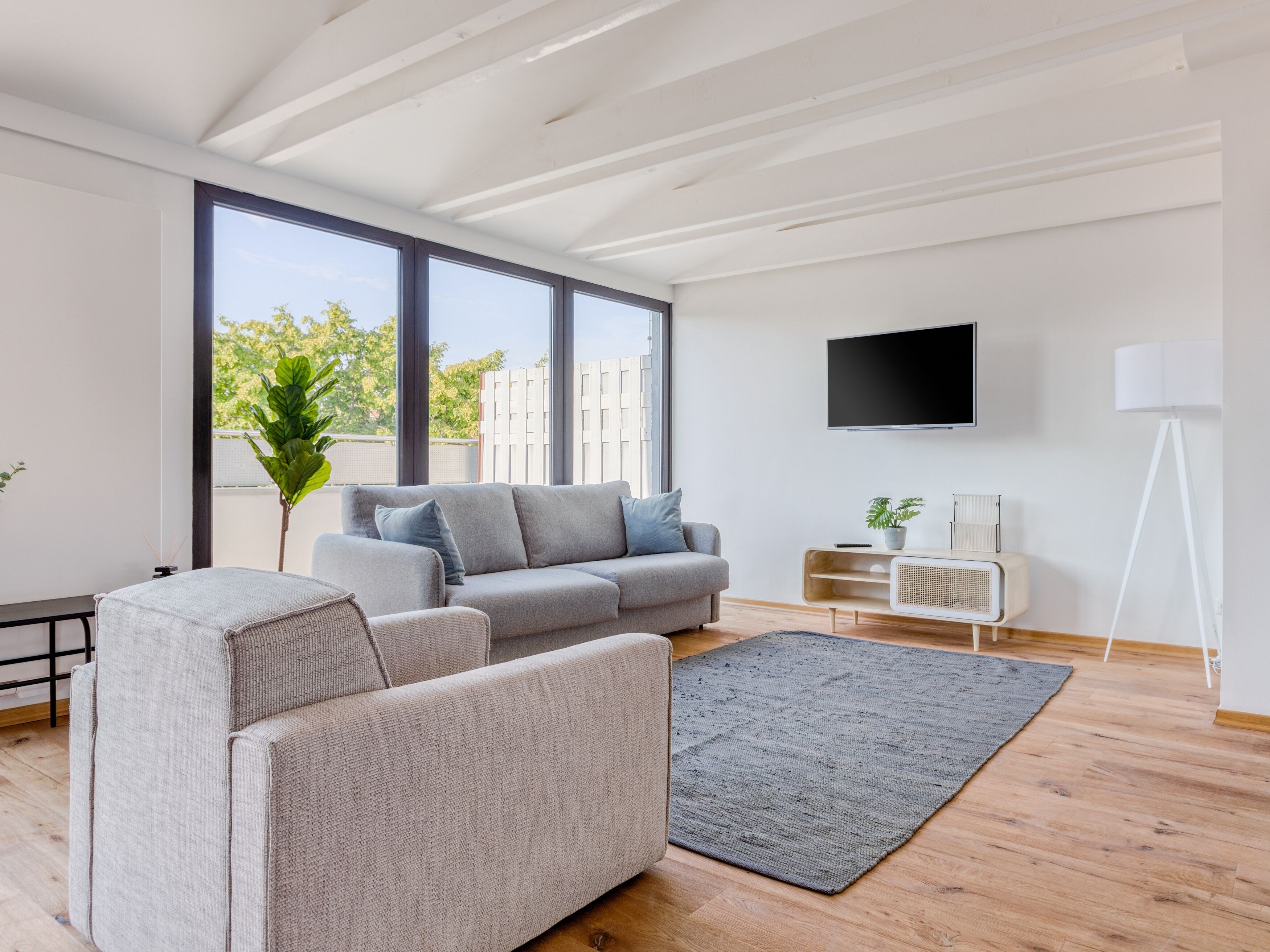 A modern living room with a grey sofa, a small table, a TV mounted on the wall, and a large window with a view of buildings and trees.