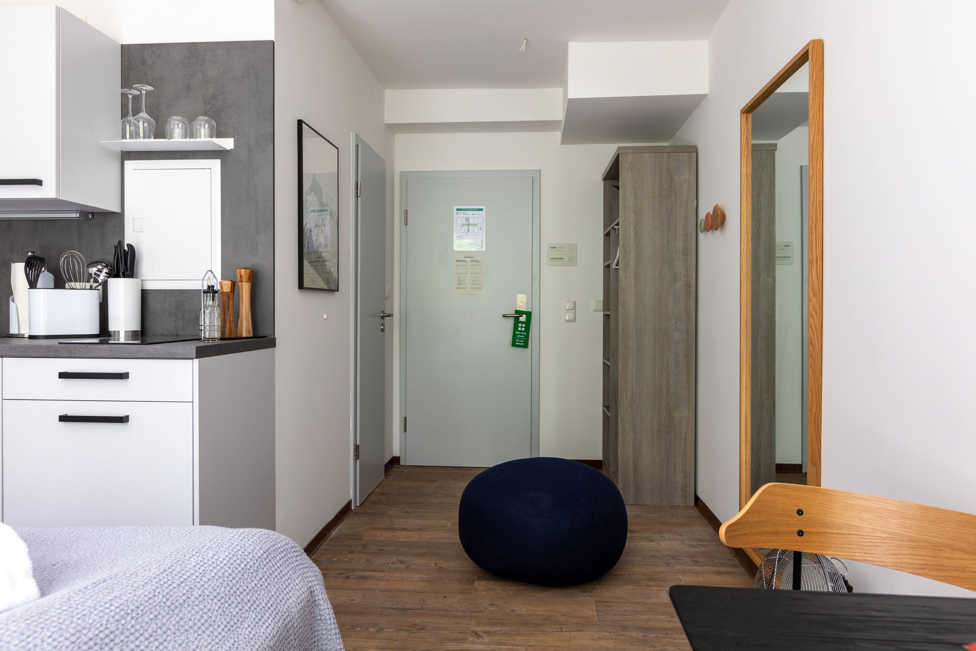 A modern hotel room with a kitchenette, a bed, a round bean bag, a wooden bench, and a large mirror.