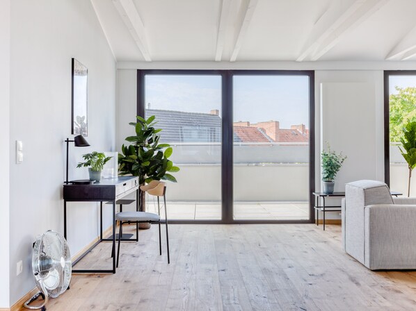 A modern living room with a large sliding glass door leading to a balcony, a desk with a chair, a sofa, and a potted plant. - Limehome Hannover Bleichenstraße (Hannover)
