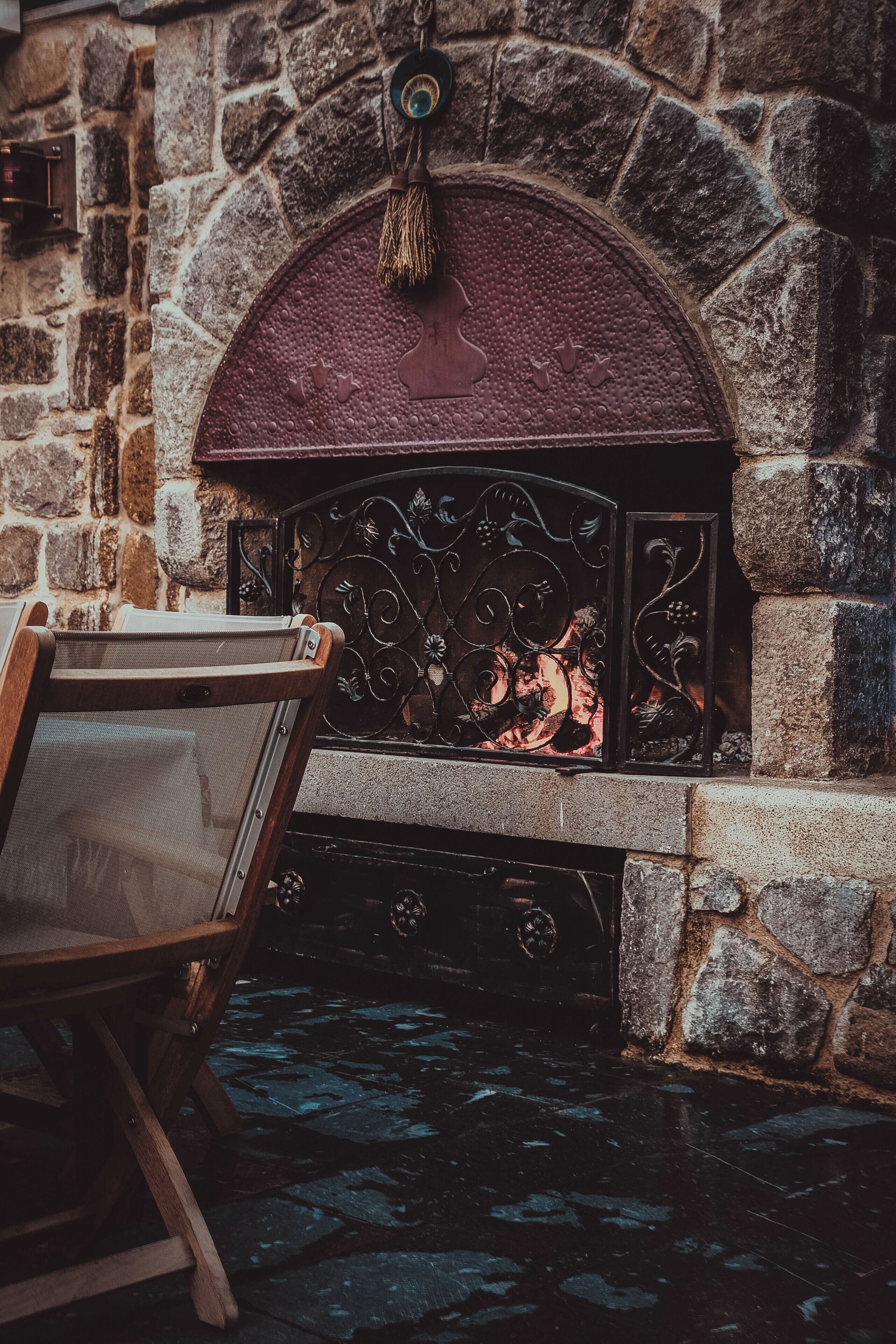 A stone fireplace with a decorative metal grate, a wooden chair in front, and a stone wall.