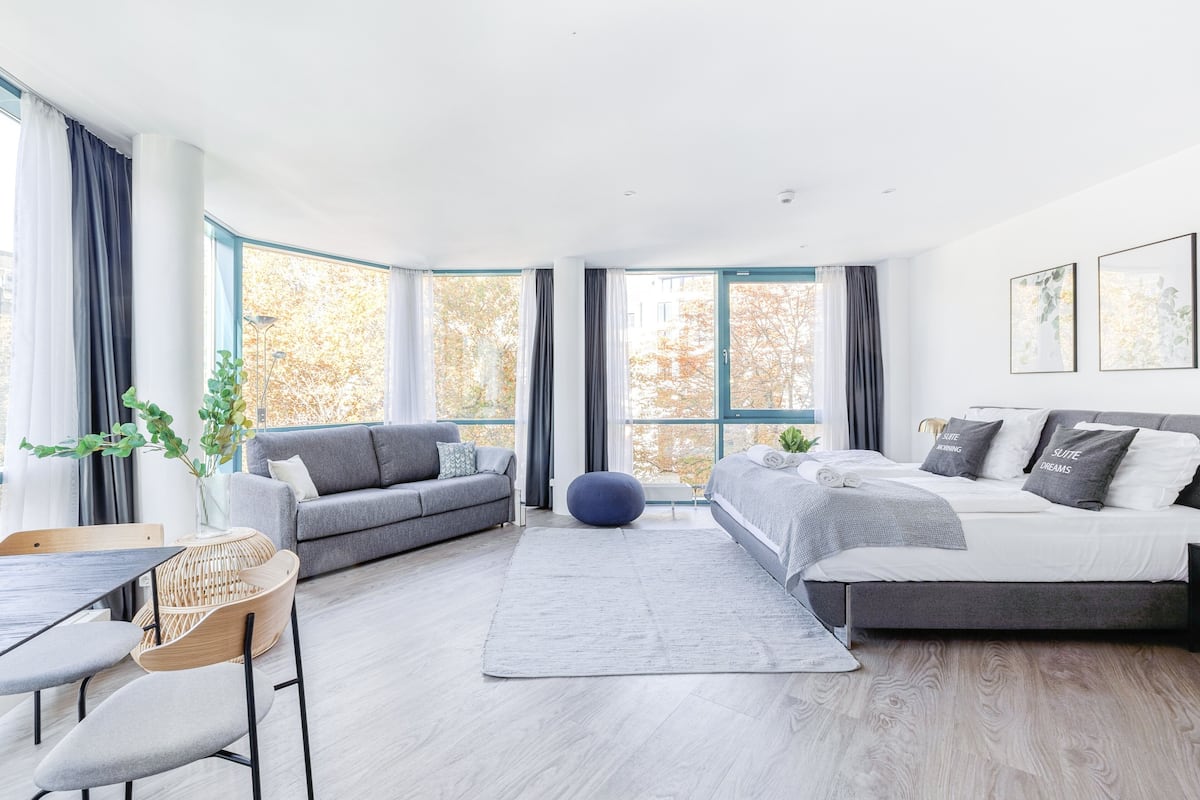 A modern, well-lit room with a sofa, a bed, a dining table, and large windows offering a view of trees.