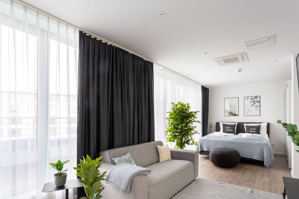 A modern bedroom with a grey sofa, a bed with grey bedding, and a large window with sheer curtains.