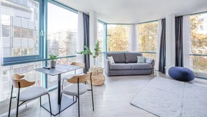 Una sala de estar moderna con un sofá gris, una mesa de comedor con sillas, un ventanal y vistas a la ciudad.