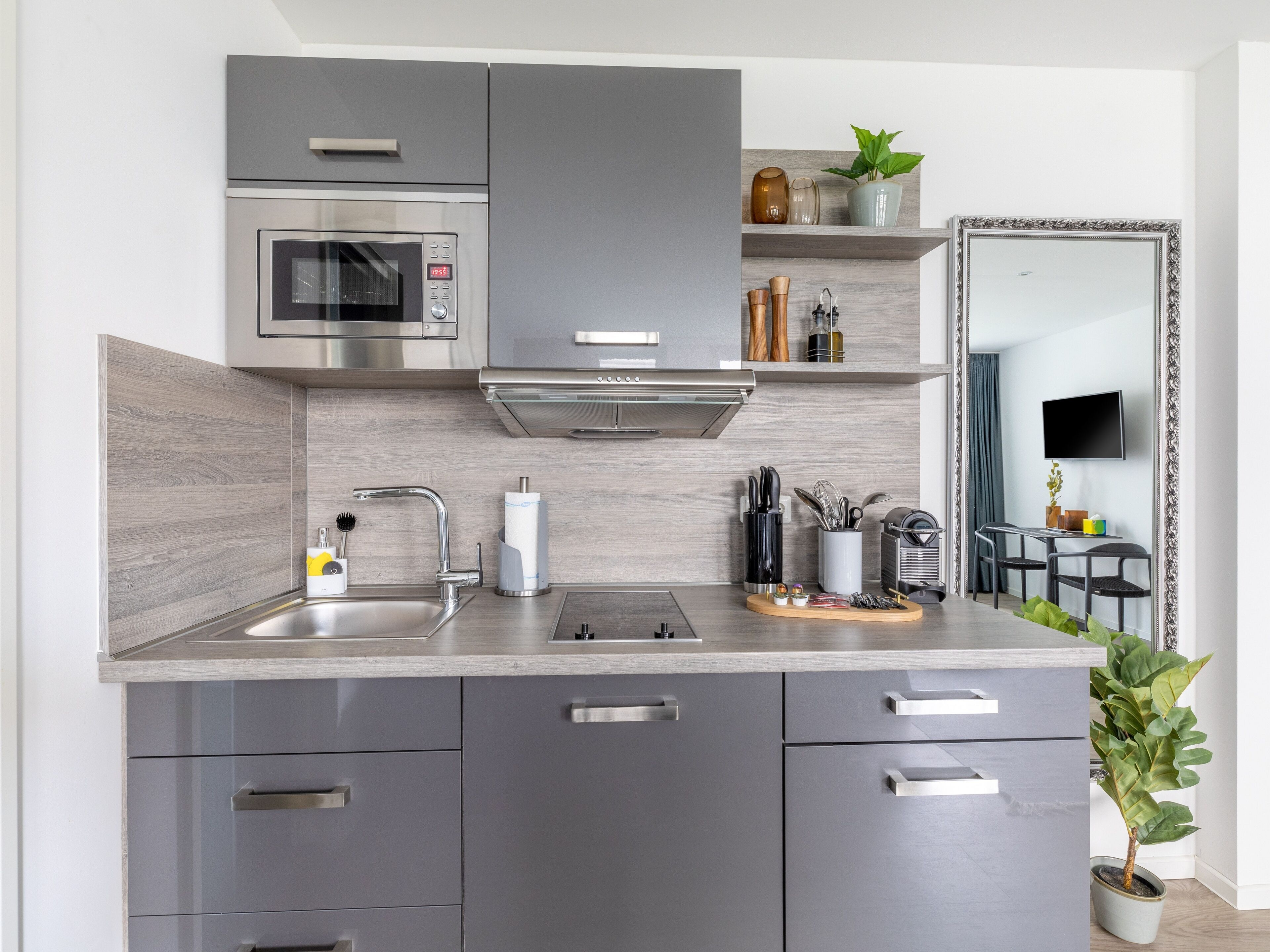 Una cocina moderna con armarios grises, un microondas de acero inoxidable y un horno empotrado. Hay repisas con adornos y una planta en maceta.