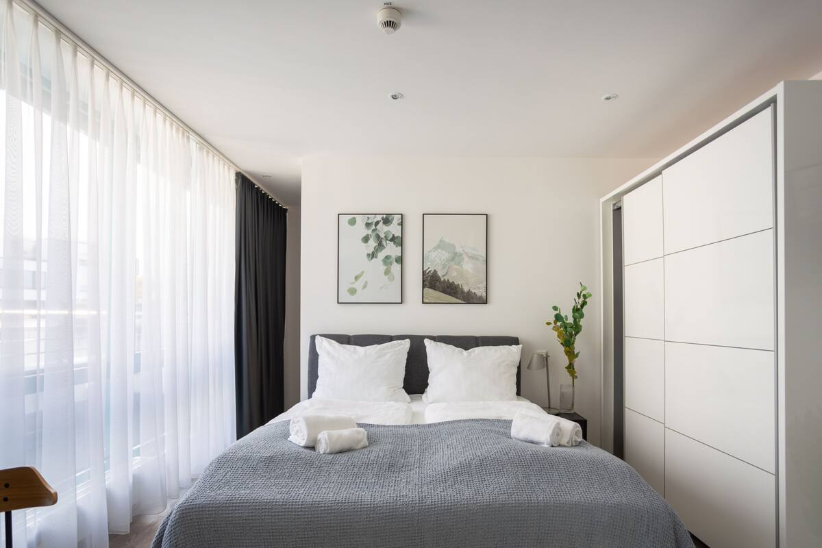 A modern hotel room with a large bed, a side table, a chair, and a wardrobe.
