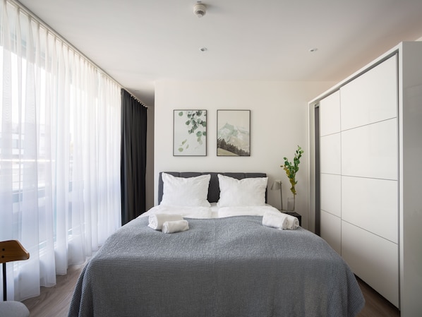 A modern hotel room with a large bed, a side table, a chair, and a wardrobe.