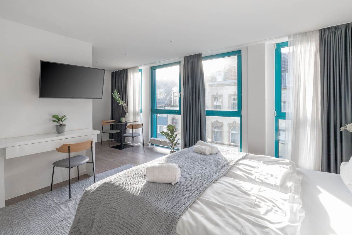 A modern hotel room with a large bed, a desk with a chair, a television, and a view of the city through large windows.
