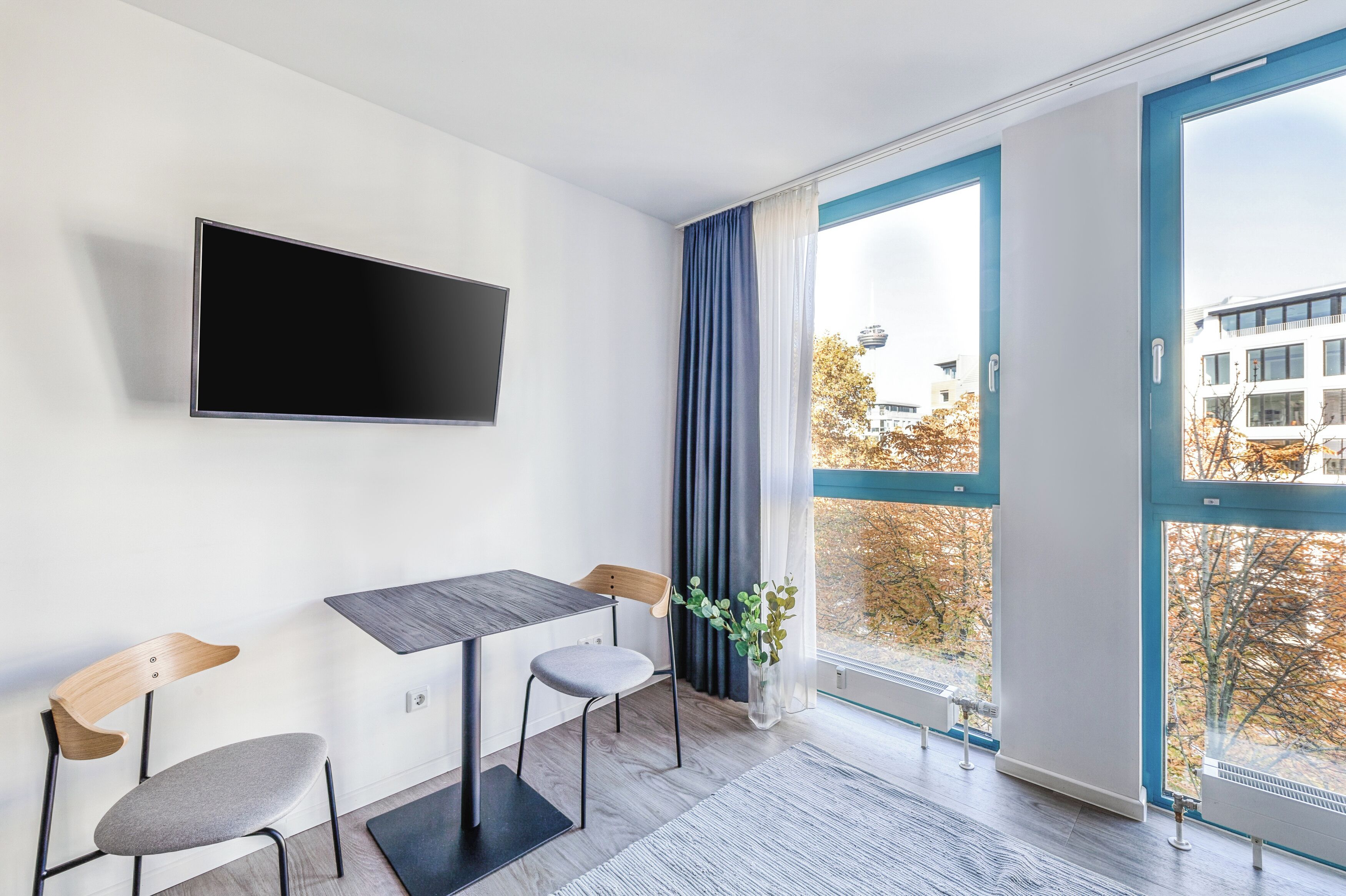 Una habitación moderna con una televisión de pantalla plana, una mesa de comedor, dos sillas, una planta en maceta y un amplio ventanal con vistas a edificios y árboles.