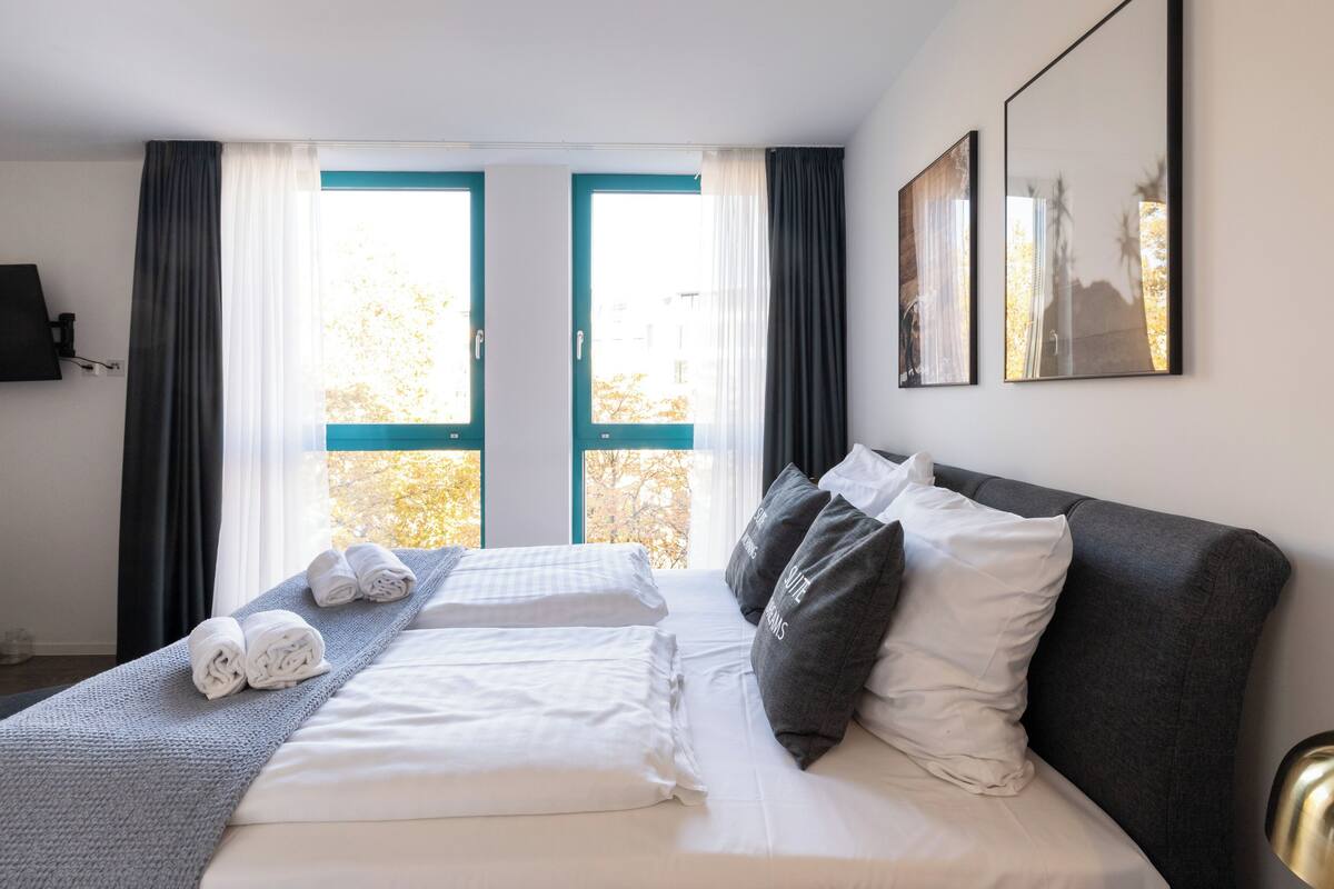 A hotel room with a bed, two windows, a painting, and a view of trees.