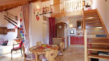 A cozy dining area with a wooden table and chairs, a kitchen in the background, and a wooden staircase.