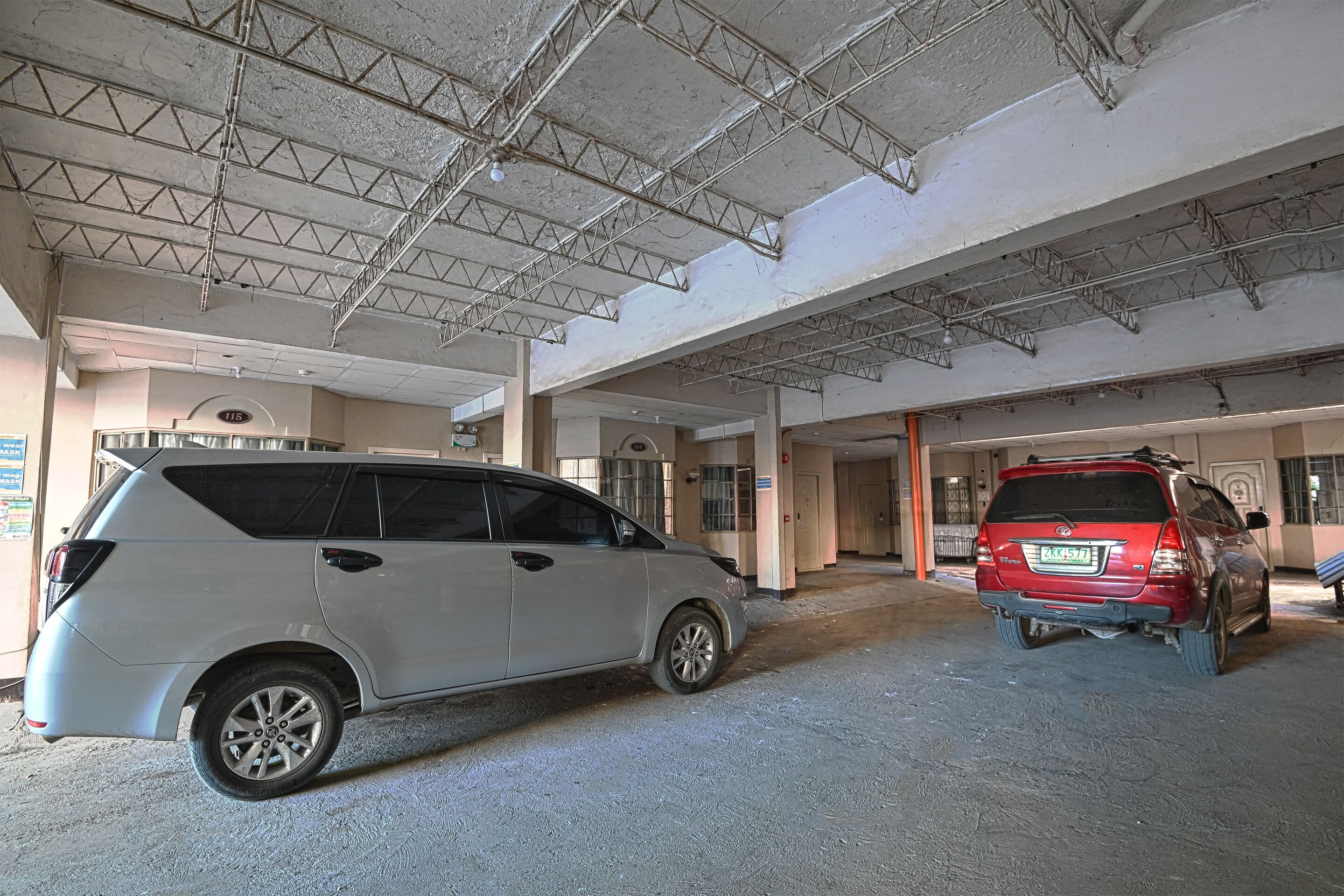 A parking area with two cars, a white SUV and a red SUV, parked inside a building with a high ceiling and visible structural beams.