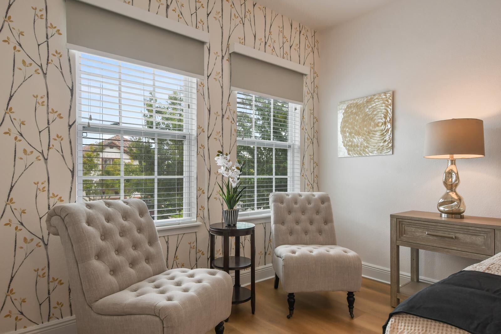 A room with two tufted armchairs, a small table with a vase of flowers, and a lamp on a wooden nightstand.