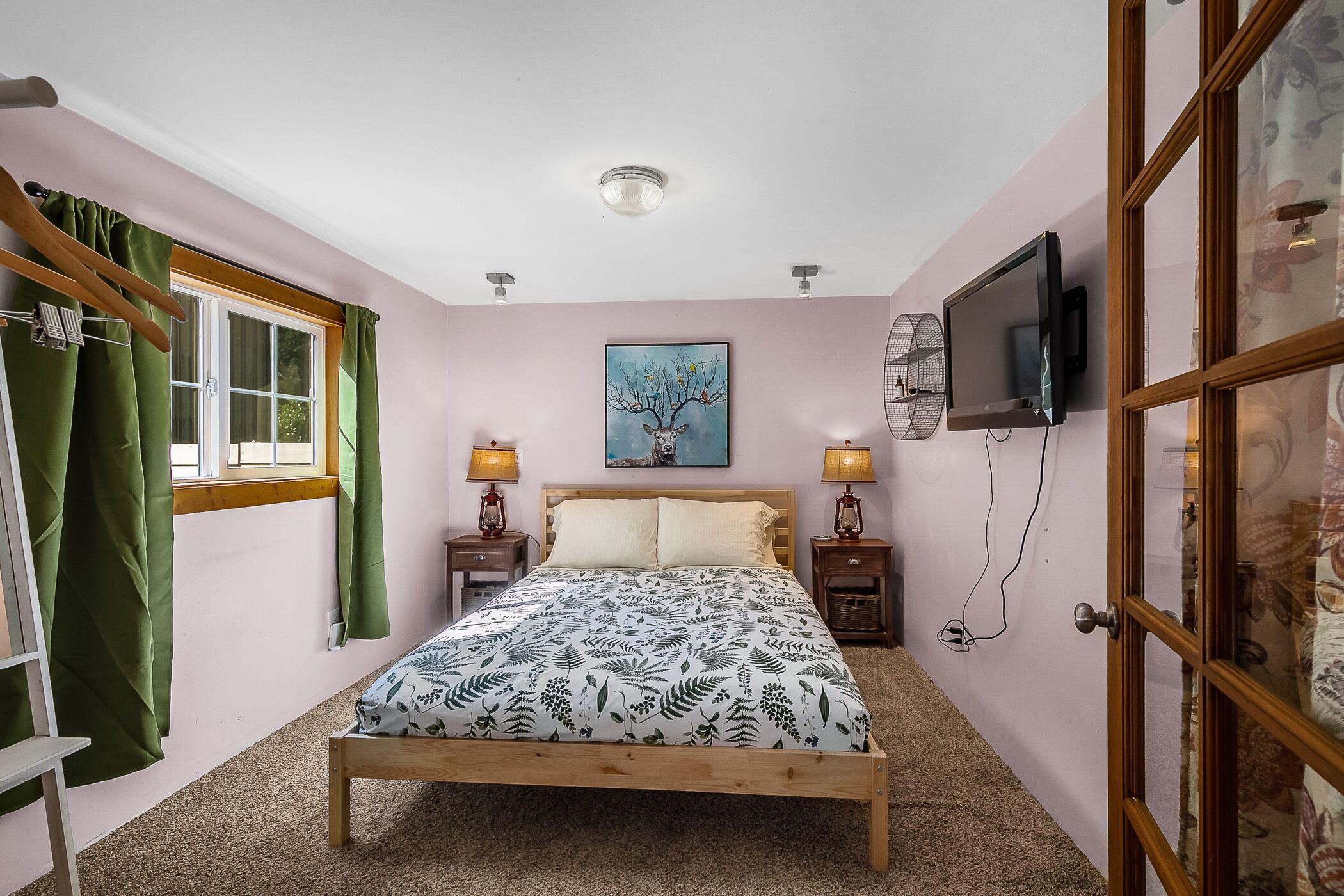A bedroom with a bed, a window with curtains, a painting on the wall, and a television on the wall.