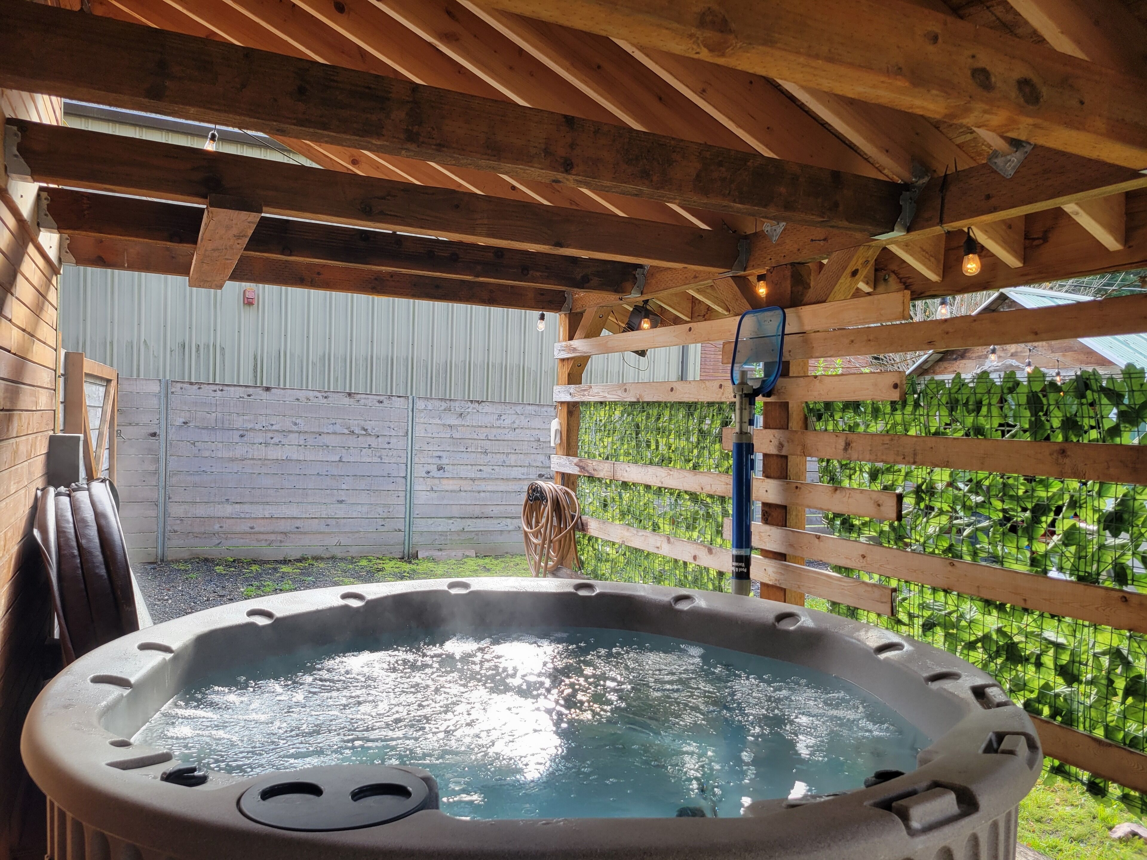 A hot tub under a wooden pergola with a metal fence and greenery in the background.