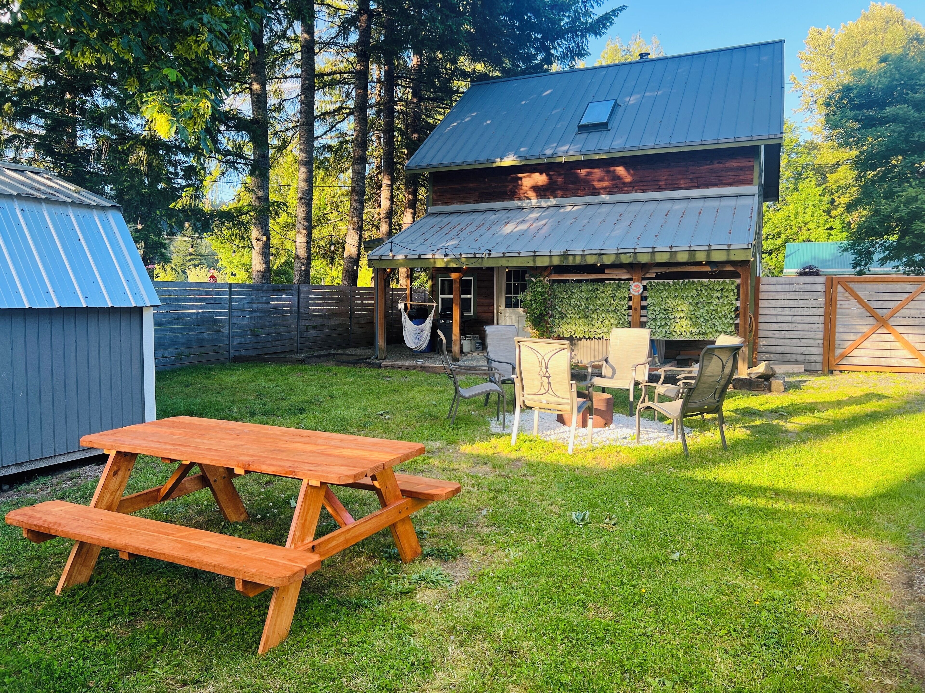 A wooden picnic table and chairs in a backyard with a covered seating area and a shed.