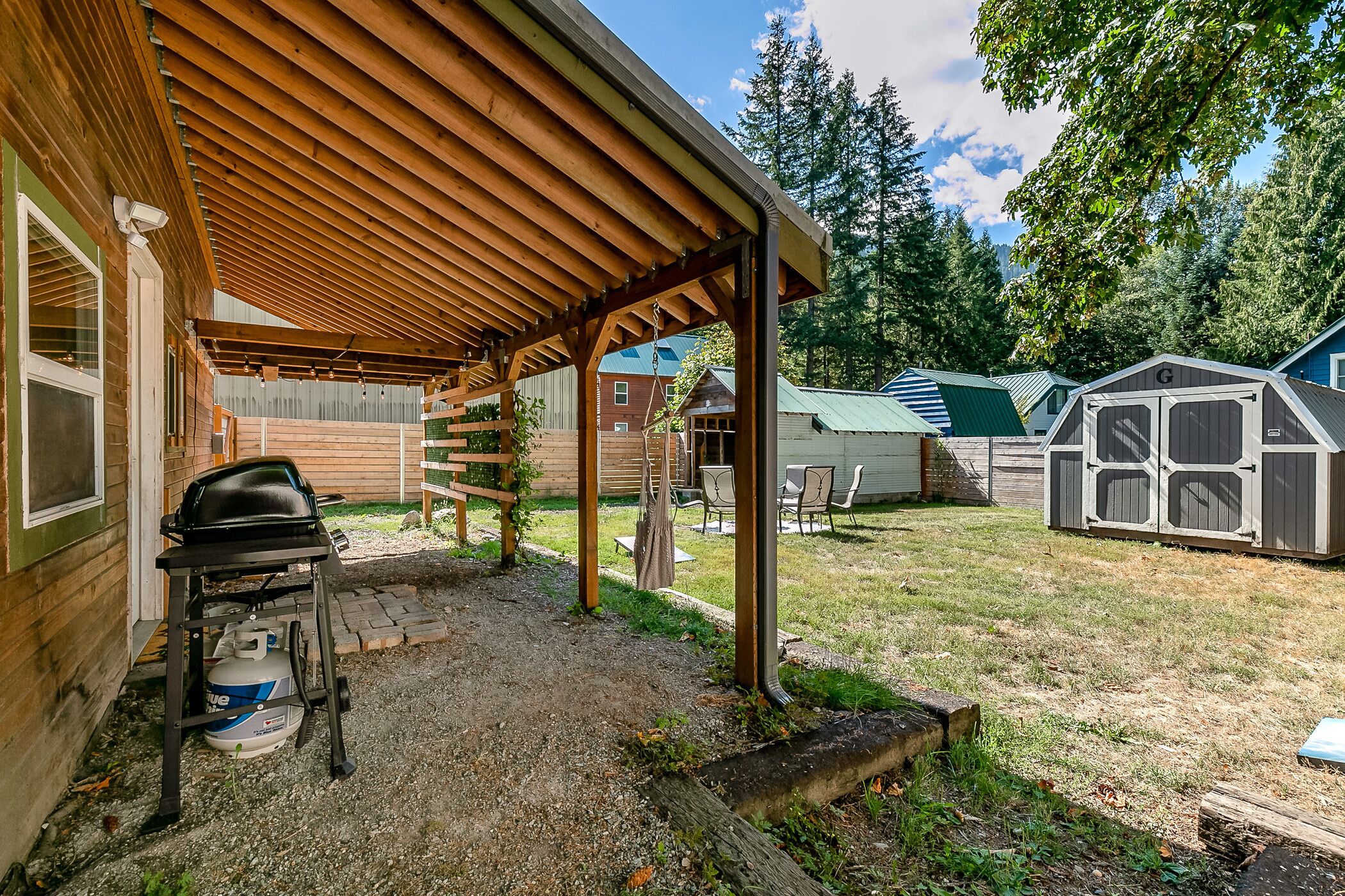 A covered patio area with a grill and propane tank, wooden deck, and a fenced yard with a shed.