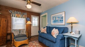 A room with a blue couch, wicker chair, wooden walls, and a ceiling fan. - Secluded Hideaway w/ Private Deep Water Dock; Sunrise Views; Kayaker's Paradise (Edisto Island)