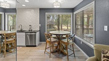 A modern kitchen with a dining area, featuring a round table, wooden chairs, a dishwasher, and a window with blinds.