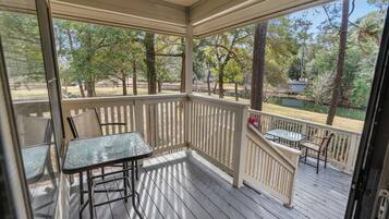 A covered outdoor deck with a glass table and chairs, surrounded by a wooden railing, overlooking a lake and trees.