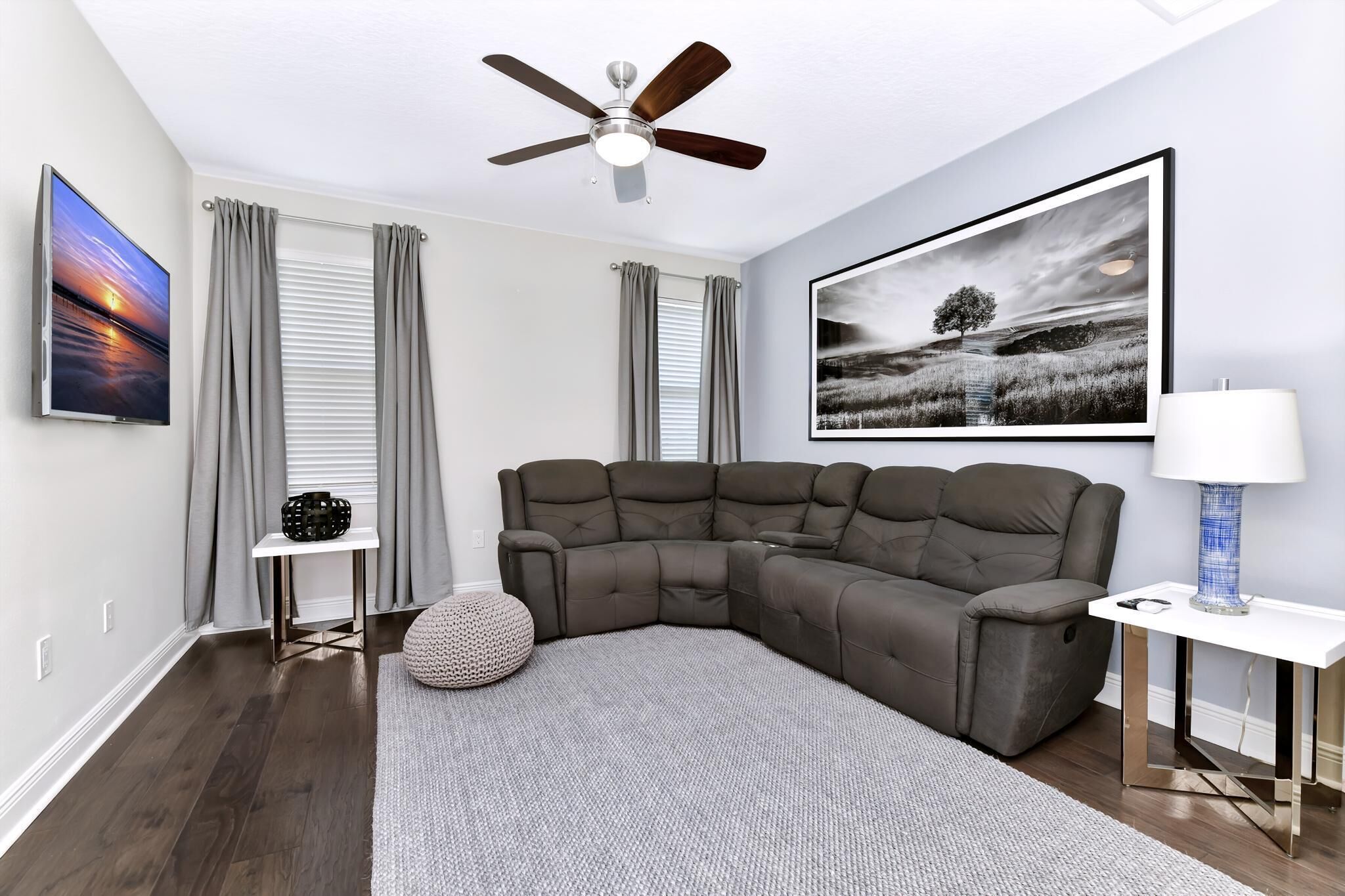 A modern living room with a sectional sofa, a ceiling fan, a framed landscape painting, a side table with a lamp, and a small decorative item on a side table.
