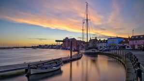 Vista frente al agua con un río tranquilo, un muelle y una embarcación llamada 'Kings Lynn', y edificios al fondo.