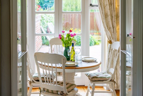A dining area with a round table, white chairs, and a vase with flowers. - Crown Villa, Wells-next-the-Sea, Norfolk (Wells-next-the-Sea)