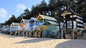 Una hilera de cabañas de playa con escaleras de madera que las conectan, sobre arena, con árboles al fondo.